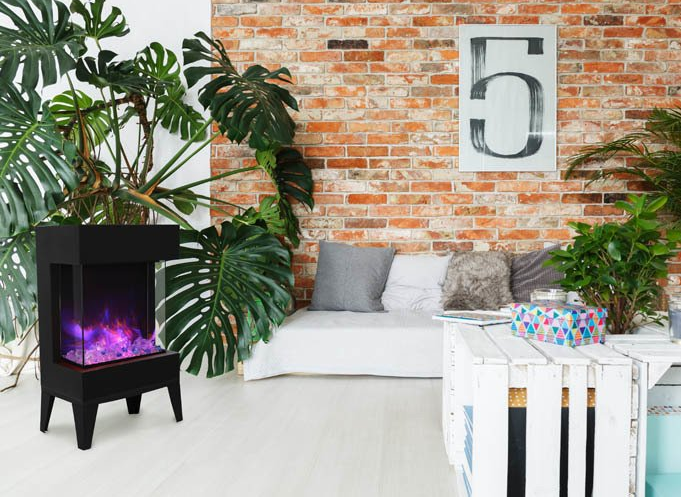 Black electric fireplace in front of plants, brick wall and white bench with pillows.