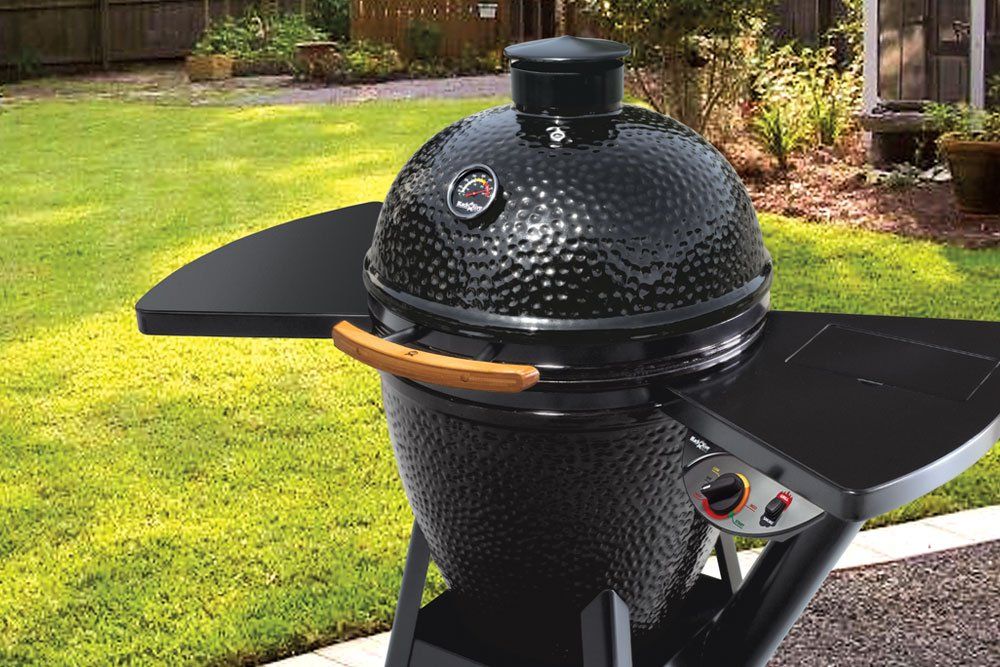 Black ceramic grill with side tables, sitting outdoors on a grassy lawn.