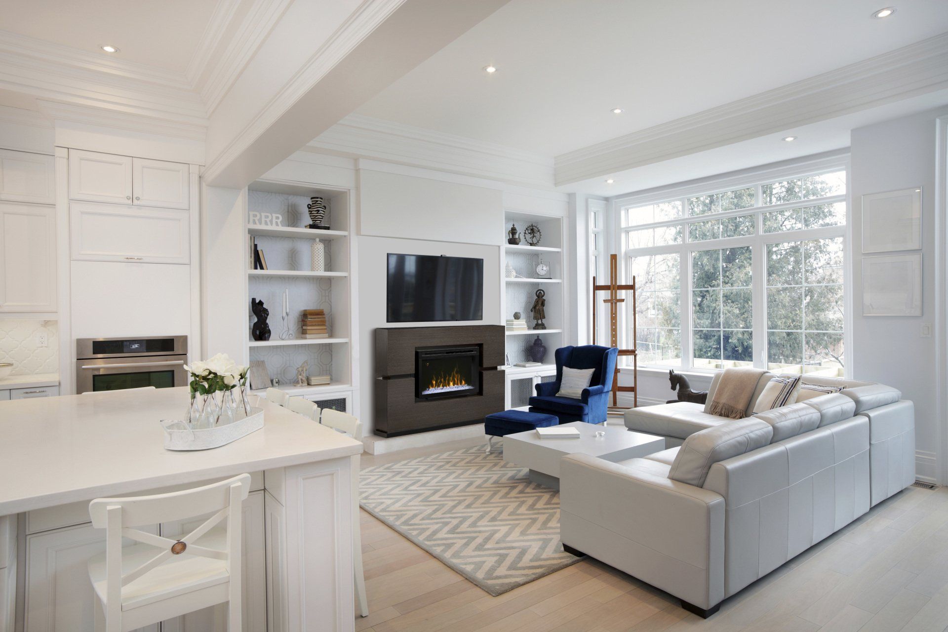 Bright, white living room with fireplace, built-in shelving, large window, sectional sofa, and kitchen island.