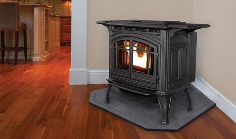 Black cast iron pellet stove with glowing fire, set on a dark gray hearth pad, in a corner of a room with hardwood floors.