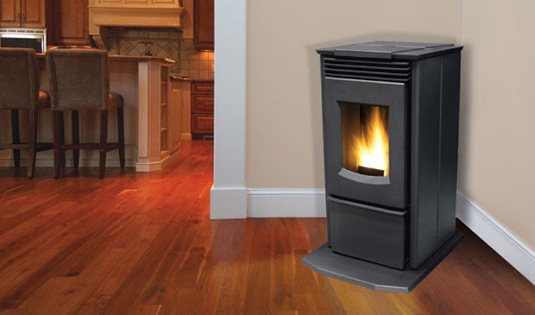Black pellet stove with flames burning in a corner of a room with wood flooring and light tan walls.