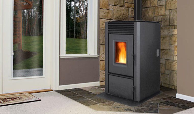 A dark gray pellet stove with a glowing fire, next to a window and stone wall.