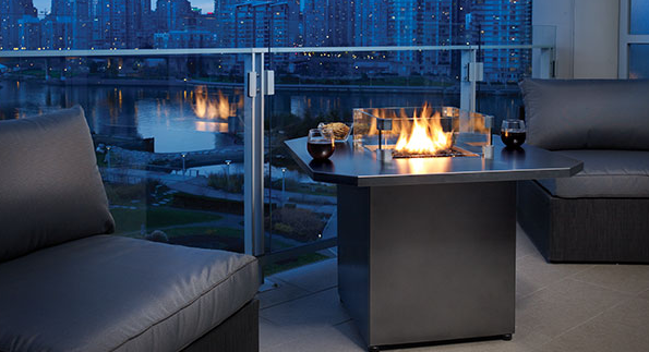 Fire pit table on a patio overlooking a city skyline at dusk; gray cushions, glass railing.
