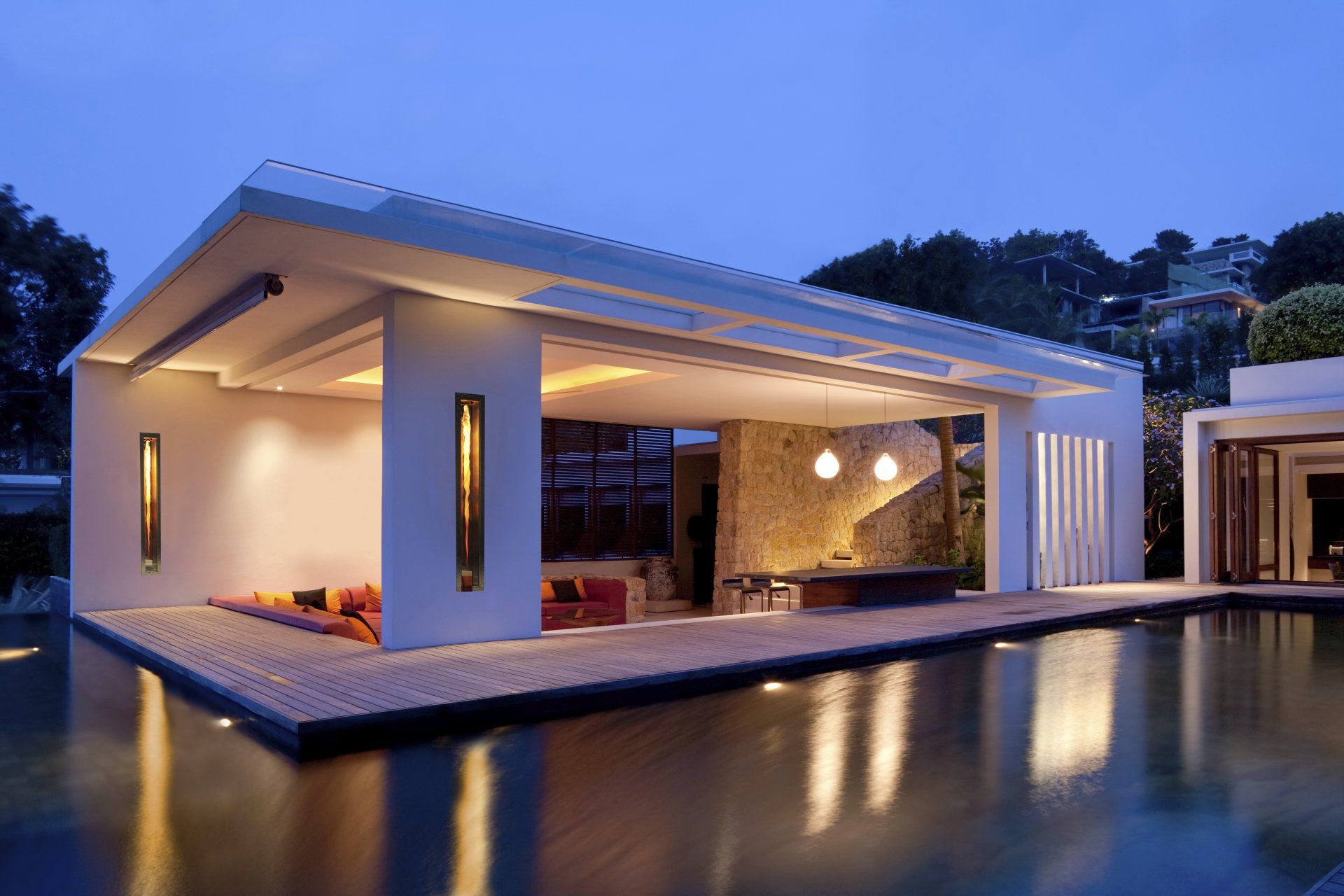 Modern illuminated poolside pavilion, open on one side, at dusk. Water reflects lights.