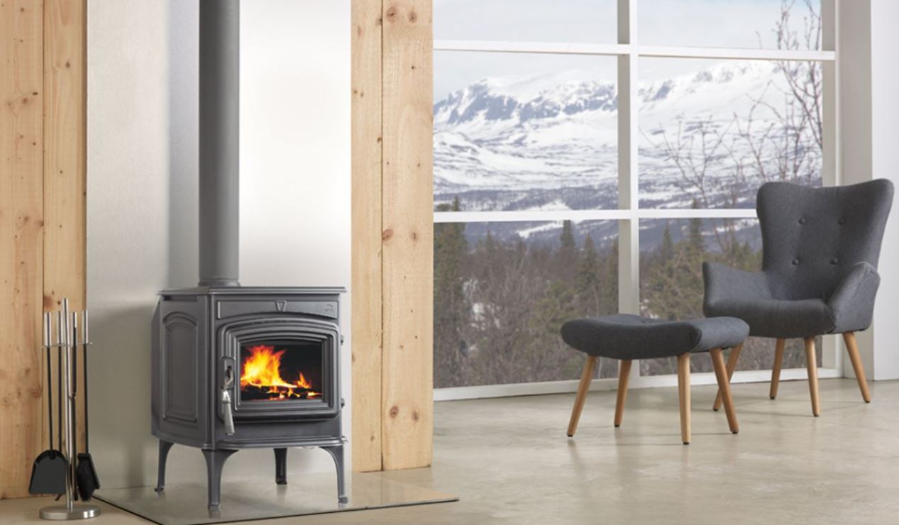 Gray wood-burning stove with flames, in a room with a window showing snowy mountains and an armchair.
