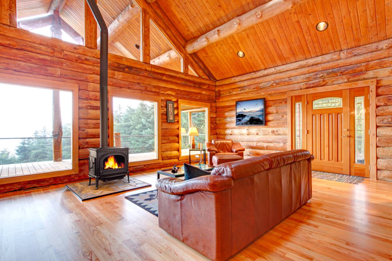 Cozy log cabin interior with wooden walls, fireplace, leather couch, and large windows.