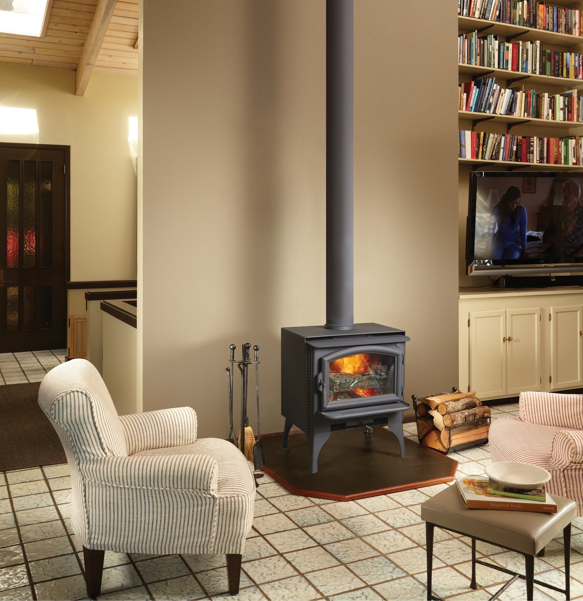 Wood-burning stove in a living room, with a chimney extending upwards. Features an armchair, tile floor, and bookshelves.