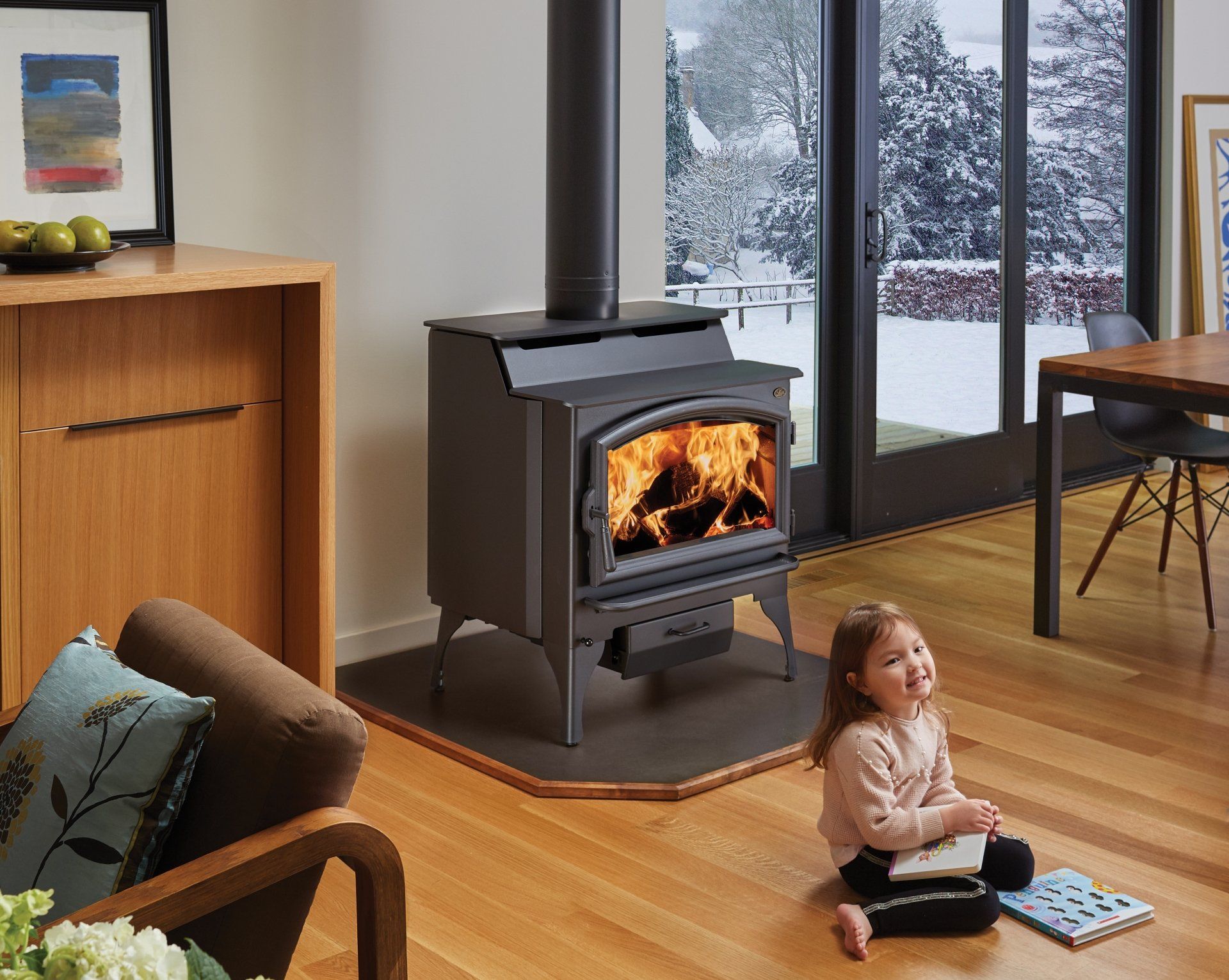 Wood-burning stove with flames, a young child on the floor, and a snowy outdoor view.