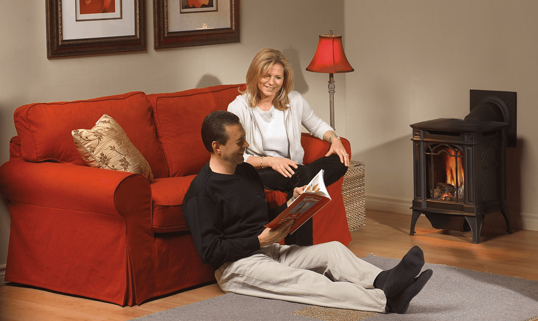 Couple in a living room; man reading on the floor, woman on the red sofa. Fireplace and lamp present.