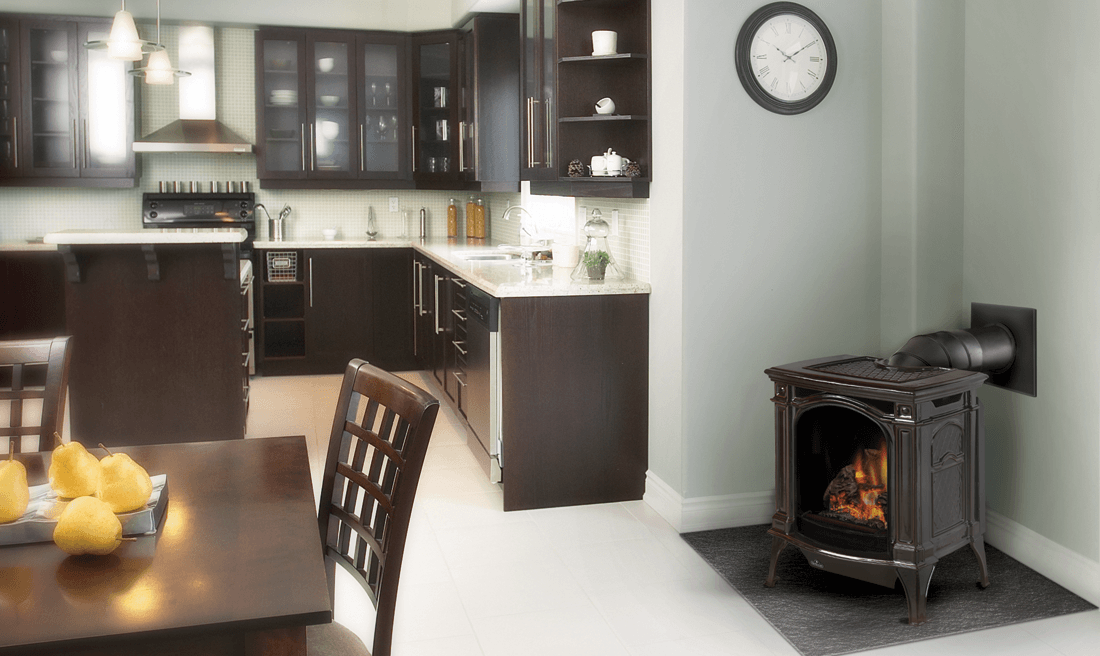A kitchen with a wood-burning stove. The stove is on a gray wall with a chimney pipe and clock. Dark wood cabinets.