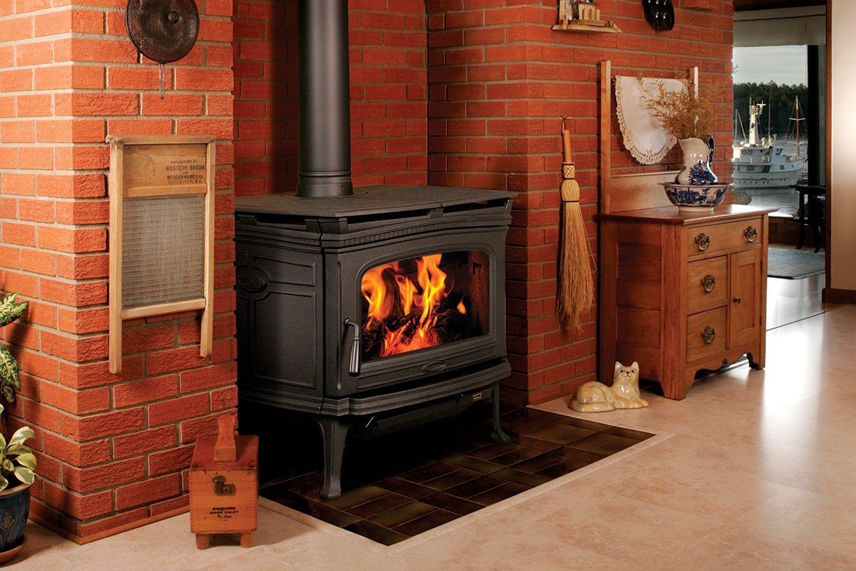 Wood-burning stove in a brick alcove with fire visible. Brown rug, wooden chest, and a laundry rack are nearby.