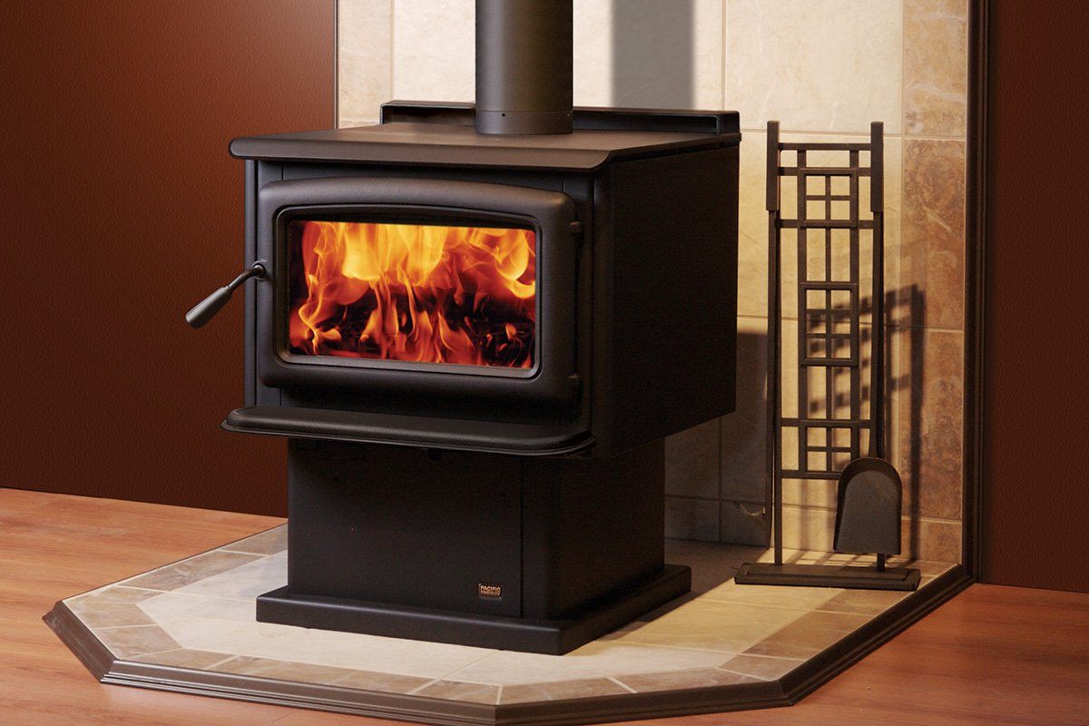 Black wood-burning stove with flames, chimney, and tool rack, in front of a stone wall.