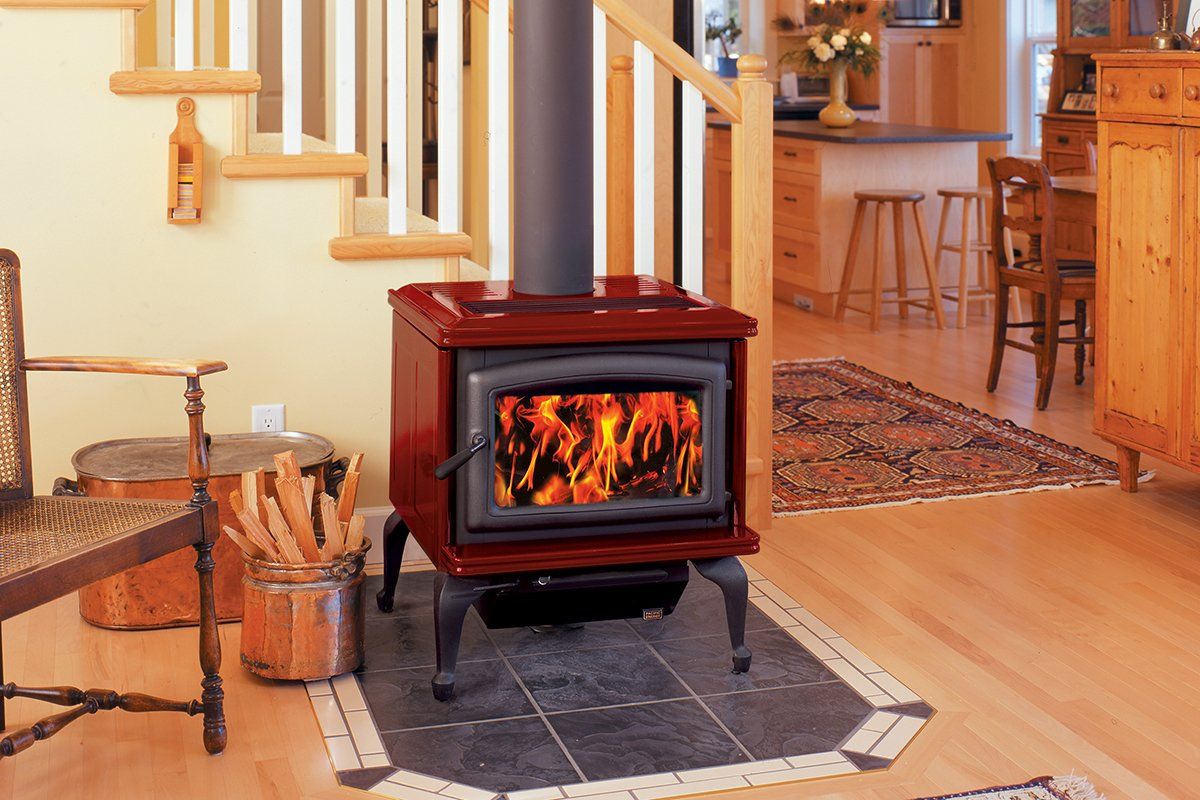 Red wood-burning stove with flames in a home; set on tile, near stairs and wooden furniture.