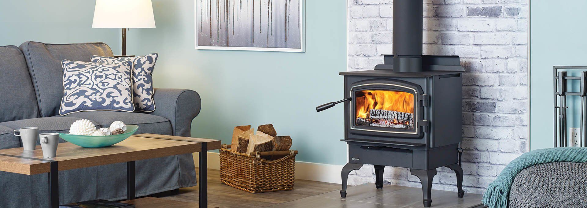 Cozy living room with a lit wood-burning stove and a grey couch. Firewood is in a wicker basket on the floor.