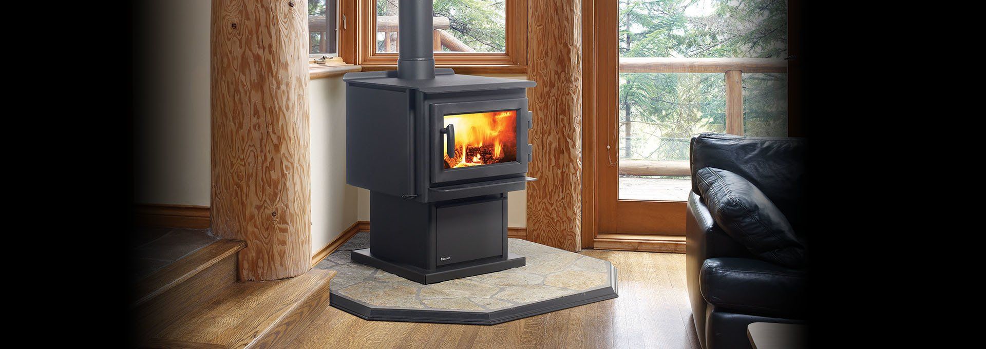 A black stove with a lit fire in front of a window and a black couch.