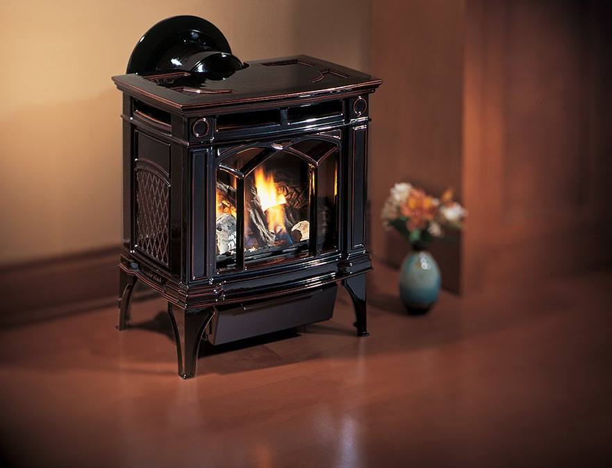 Black gas stove with a glowing fire, indoors on a wood floor near a vase of flowers.