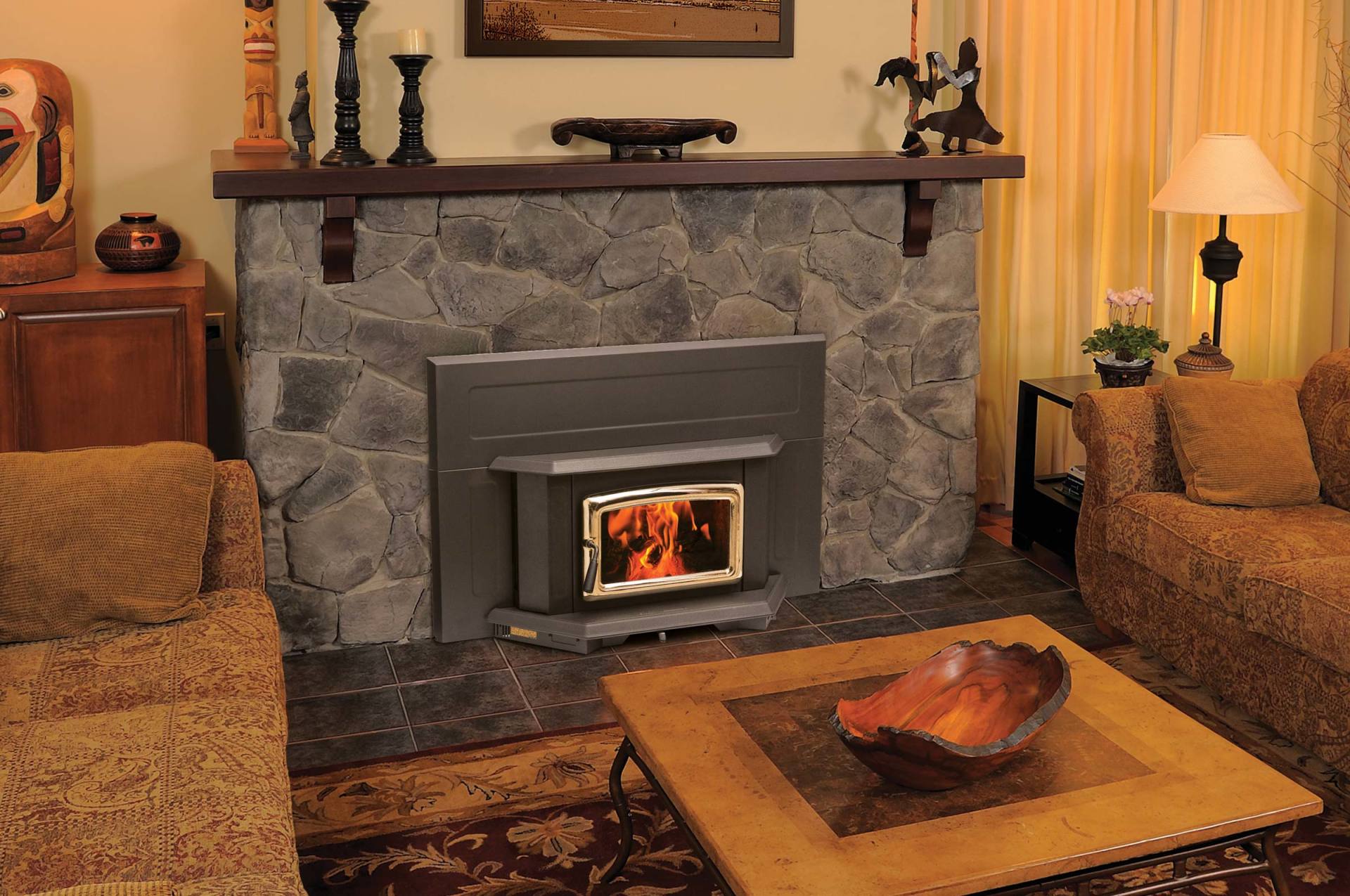 Fireplace with stone facade, dark mantel, lit fire, flanked by couches, lamp, wooden coffee table.