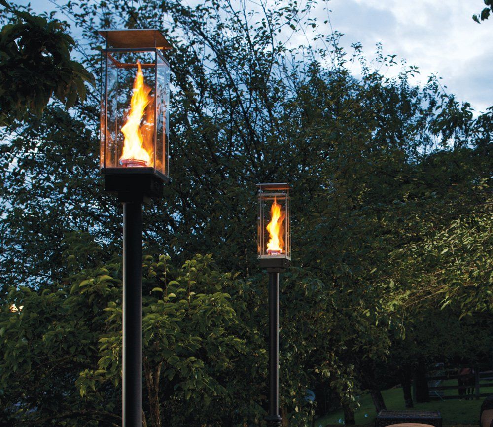 Two outdoor gas lanterns with enclosed flames on black poles, set in a lush green garden.