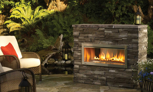 Outdoor stone fireplace with flames, next to wicker seating in a garden setting.