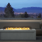 Outdoor fire pit with flames, concrete base, and couch, overlooking a city and mountains at dusk.