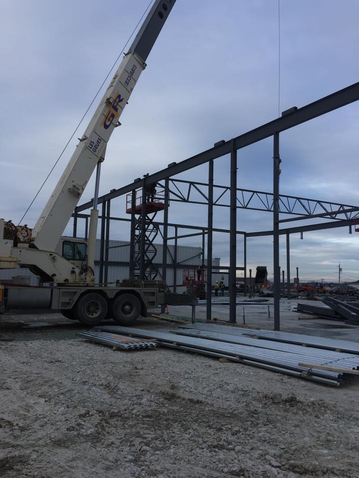 Grue soulevant des poutres d'acier sur un chantier de construction à côté d'une charpente métallique partiellement édifiée.