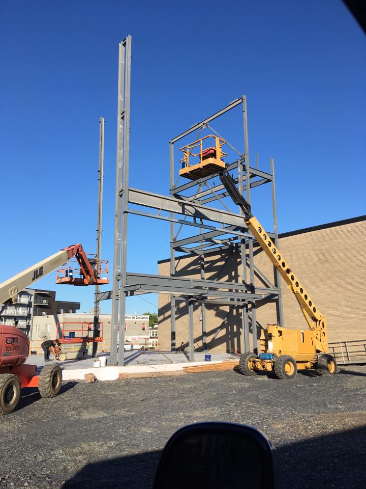 Chantier de construction avec structure métallique, nacelle jaune et ouvriers sur échafaudage sous un ciel bleu