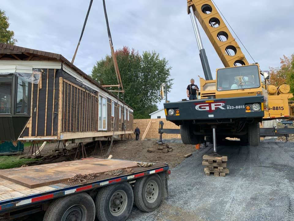 Une grue soulève un wagon ancien sur une remorque à plateau sur un chantier de gravier.