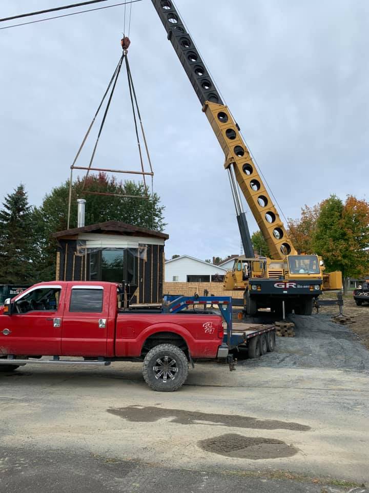 Une grue soulève un petit abri sur une remorque à côté d'une camionnette rouge sur un chantier de construction