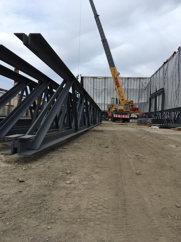 Chantier de construction avec une grue soulevant des poutres d'acier à côté de matériaux empilés sur un chemin de terre