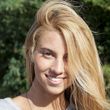 Una persona sorridente con lunghi capelli biondi si trova all'aperto, sullo sfondo di una vegetazione verde.