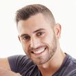 Una persona sorridente con capelli corti e scuri, barba curata e un piccolo orecchino a perno, in posa su uno sfondo bianco.