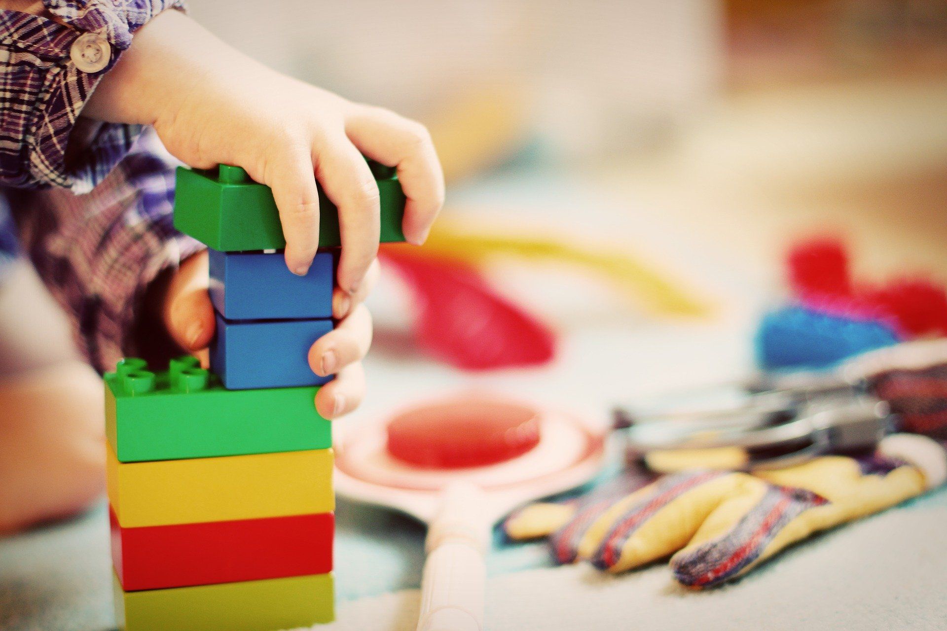 Child's hand stacking colorful toy blocks. Playroom setting with other toys.