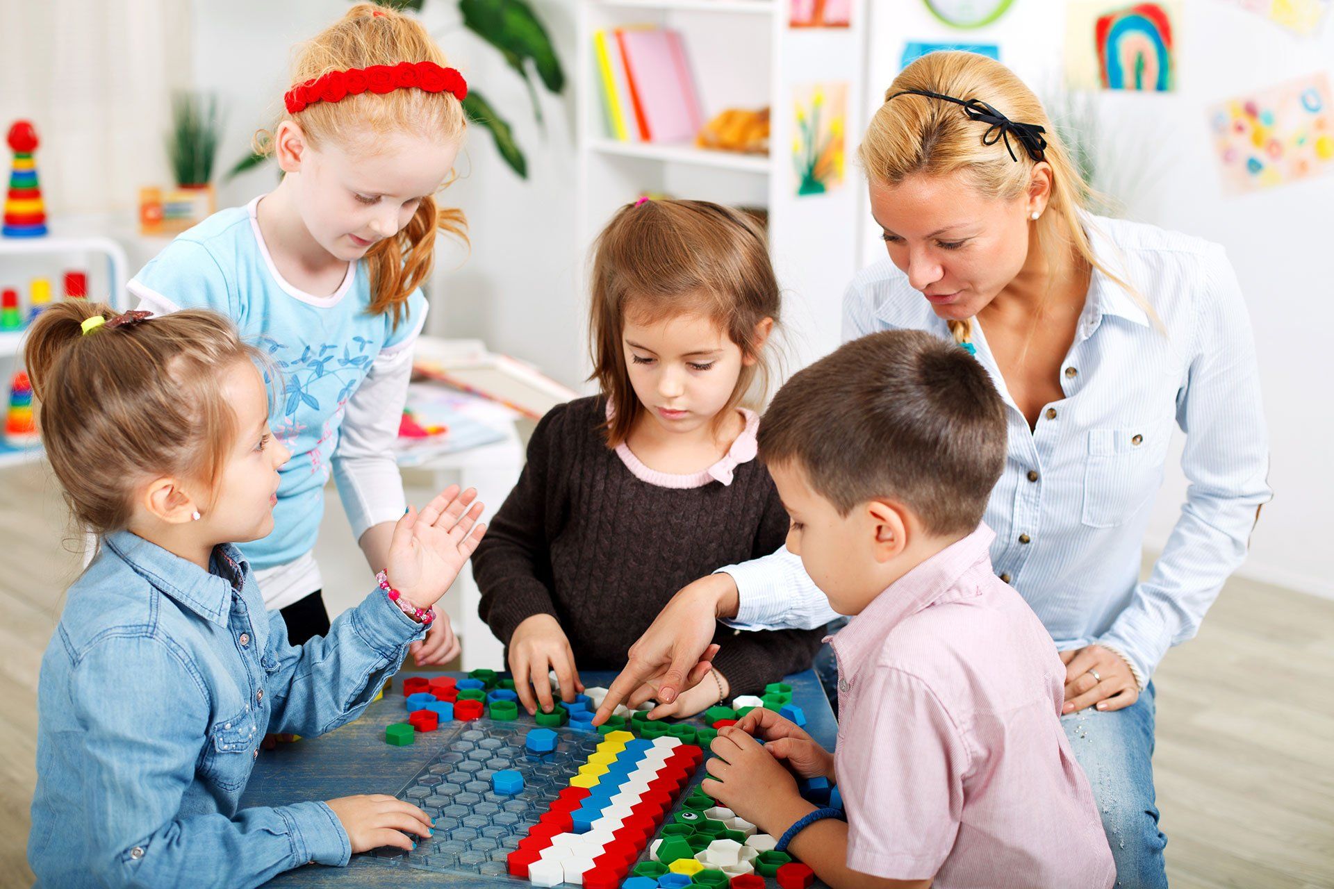 Children and a teacher play with colorful blocks on the floor in a bright classroom.