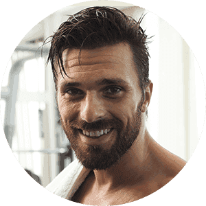 A man with a beard is smiling in a gym with a towel around his neck.