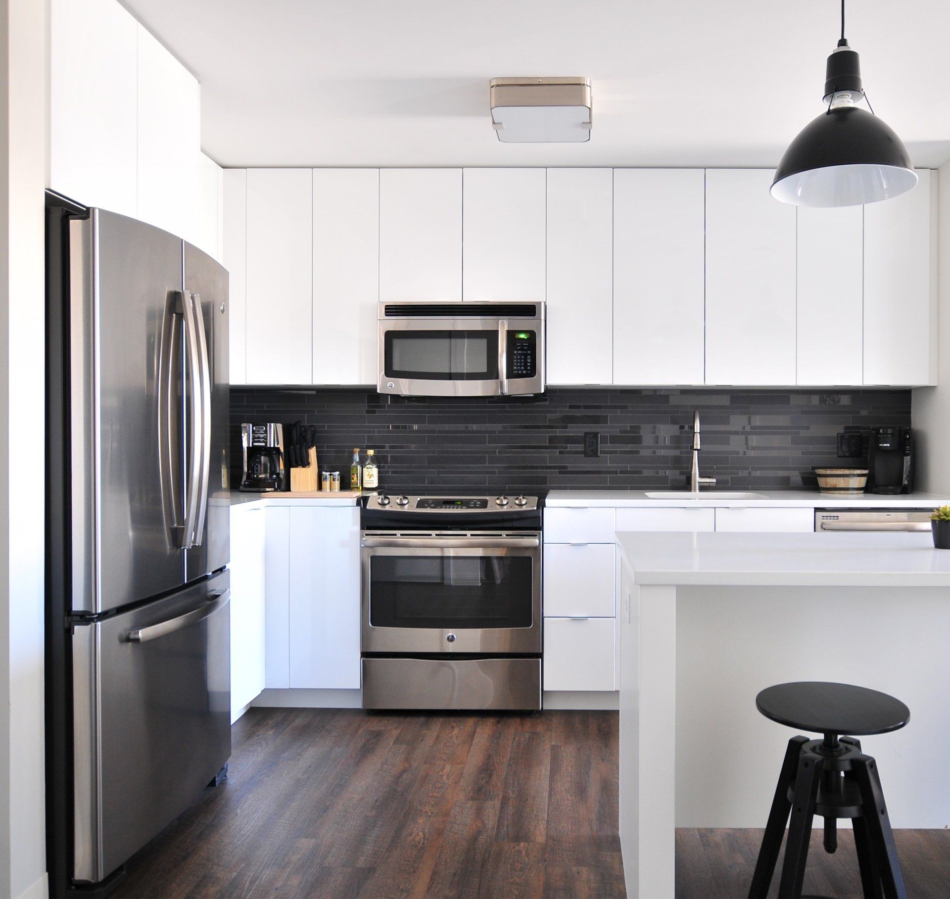 Modern white kitchen with stainless steel appliances, dark wood floor, and black pendant light