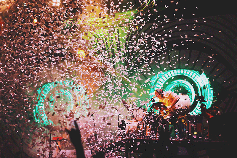 A musician performs on stage amidst a dense shower of confetti, backlit by glowing circular lights in a dark venue.