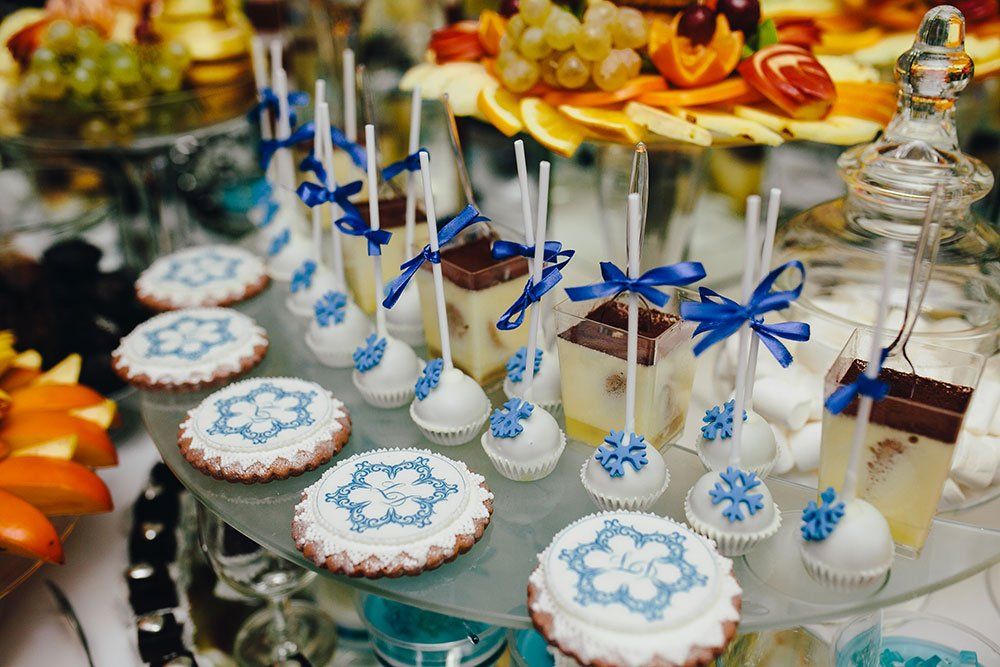 A dessert display featuring decorated snowflake cookies, cake pops with blue bows, and small layered cakes on a glass tray.