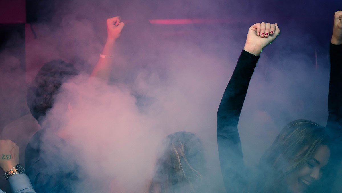 People dance in a dimly lit, smoke-filled club, raising their hands in celebration under moody purple lighting.
