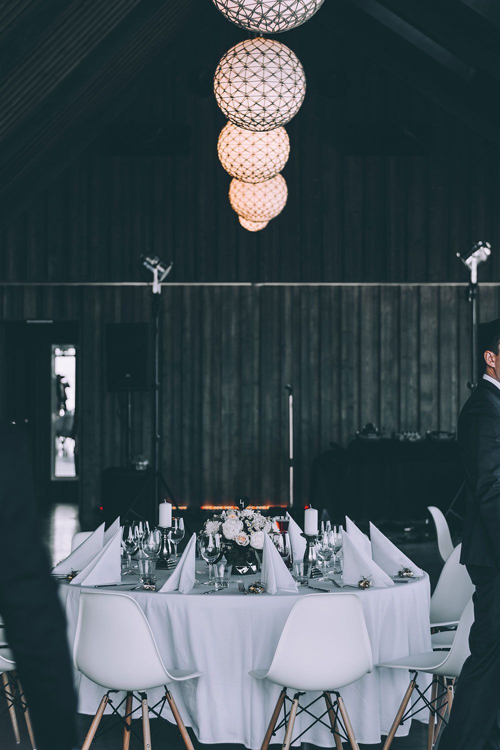 A formal dinner table with white tablecloth and chairs, set under four glowing spherical pendant lights in a dark room.