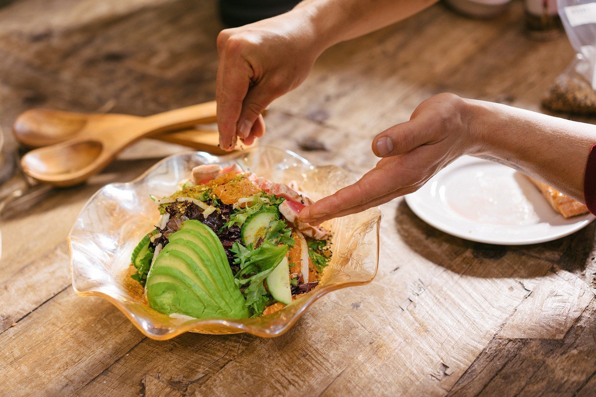 Une personne ajoute de l'avocat à une salade sur une table en bois.