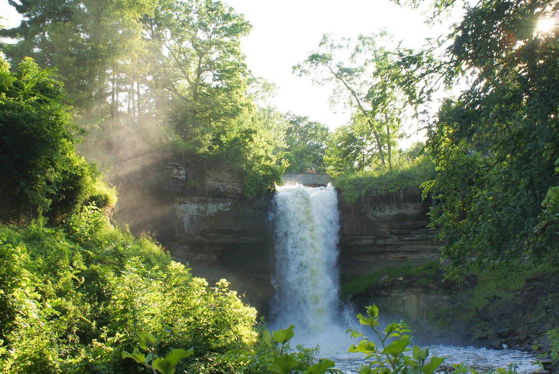 A waterfall in the middle of a forest with the sun shining through the trees