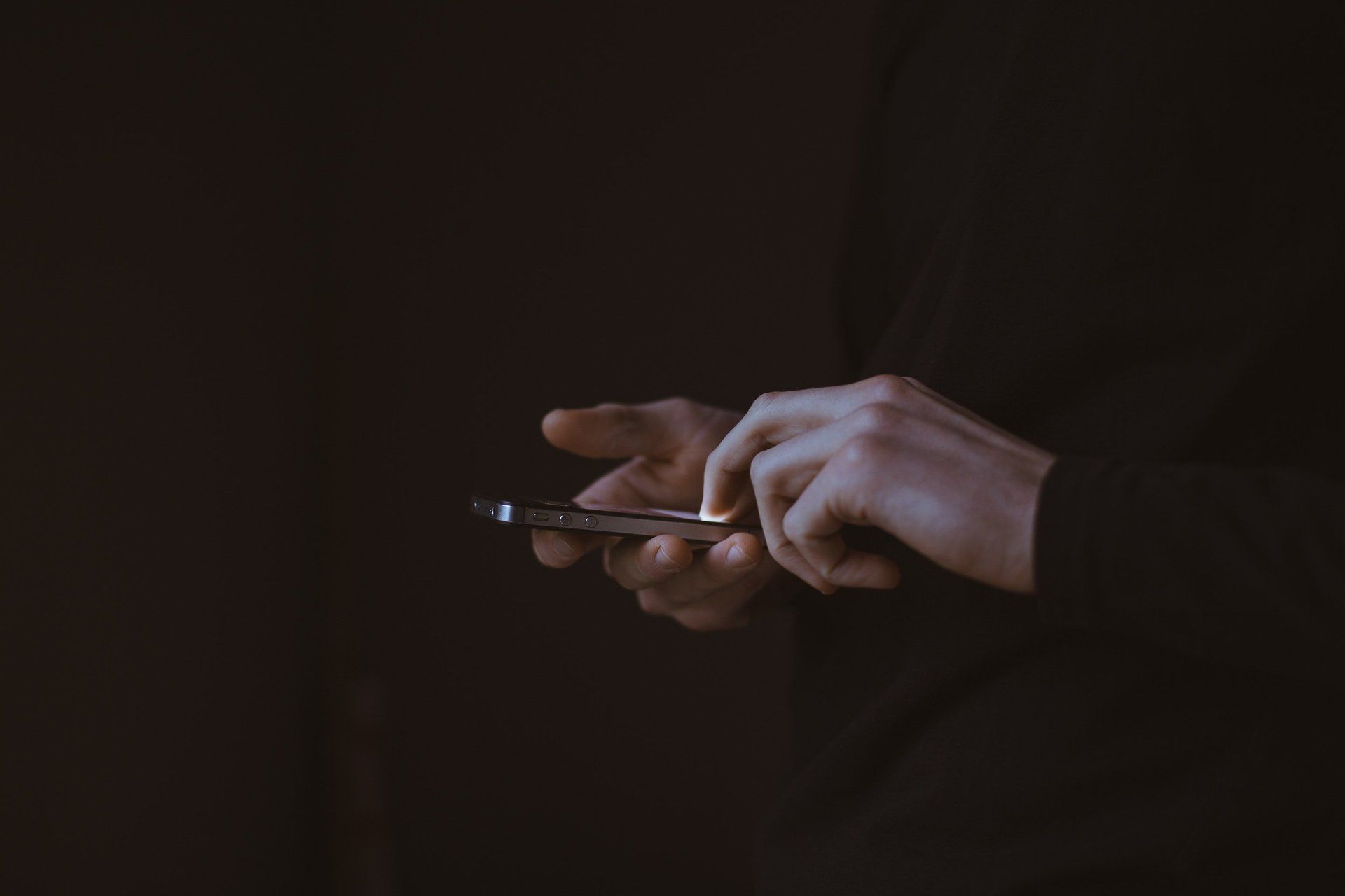 A person in a black long-sleeved shirt holds a smartphone, using one finger to tap or swipe on the screen in low light.