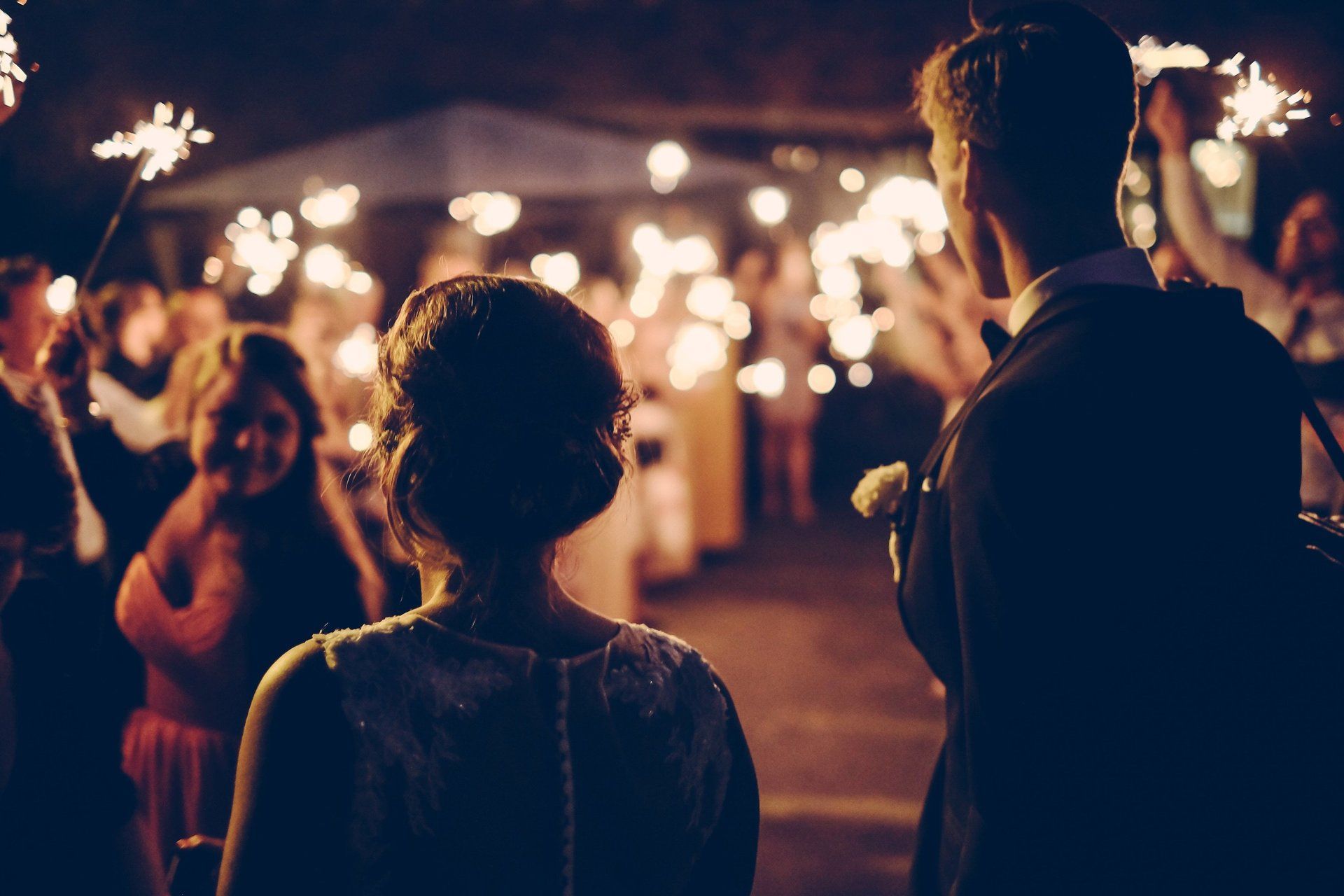 Bride and groom walk through a sparkler-lined path after a wedding, night setting.