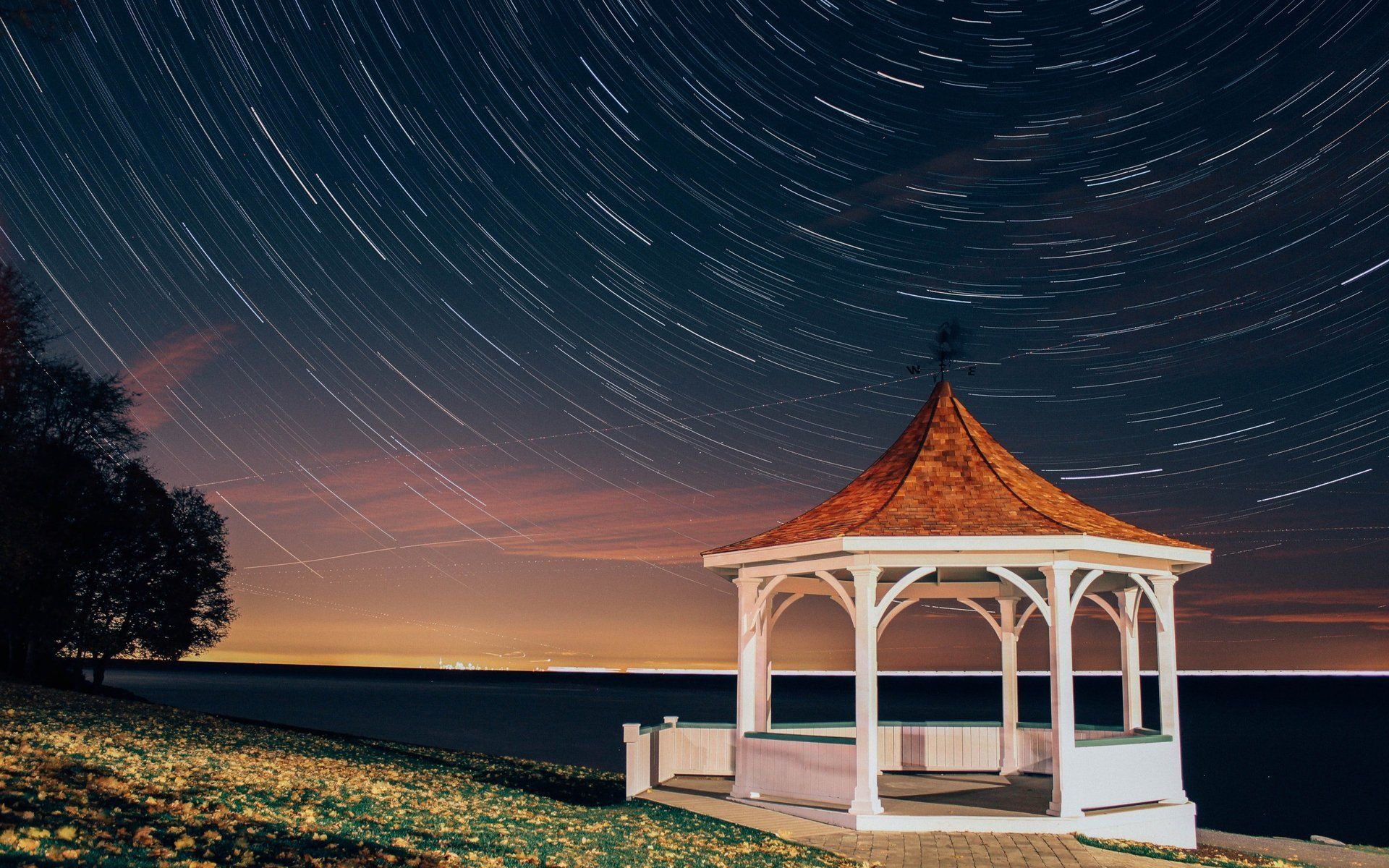 A gazebo on the shore of a lake at night with star trails in the sky.