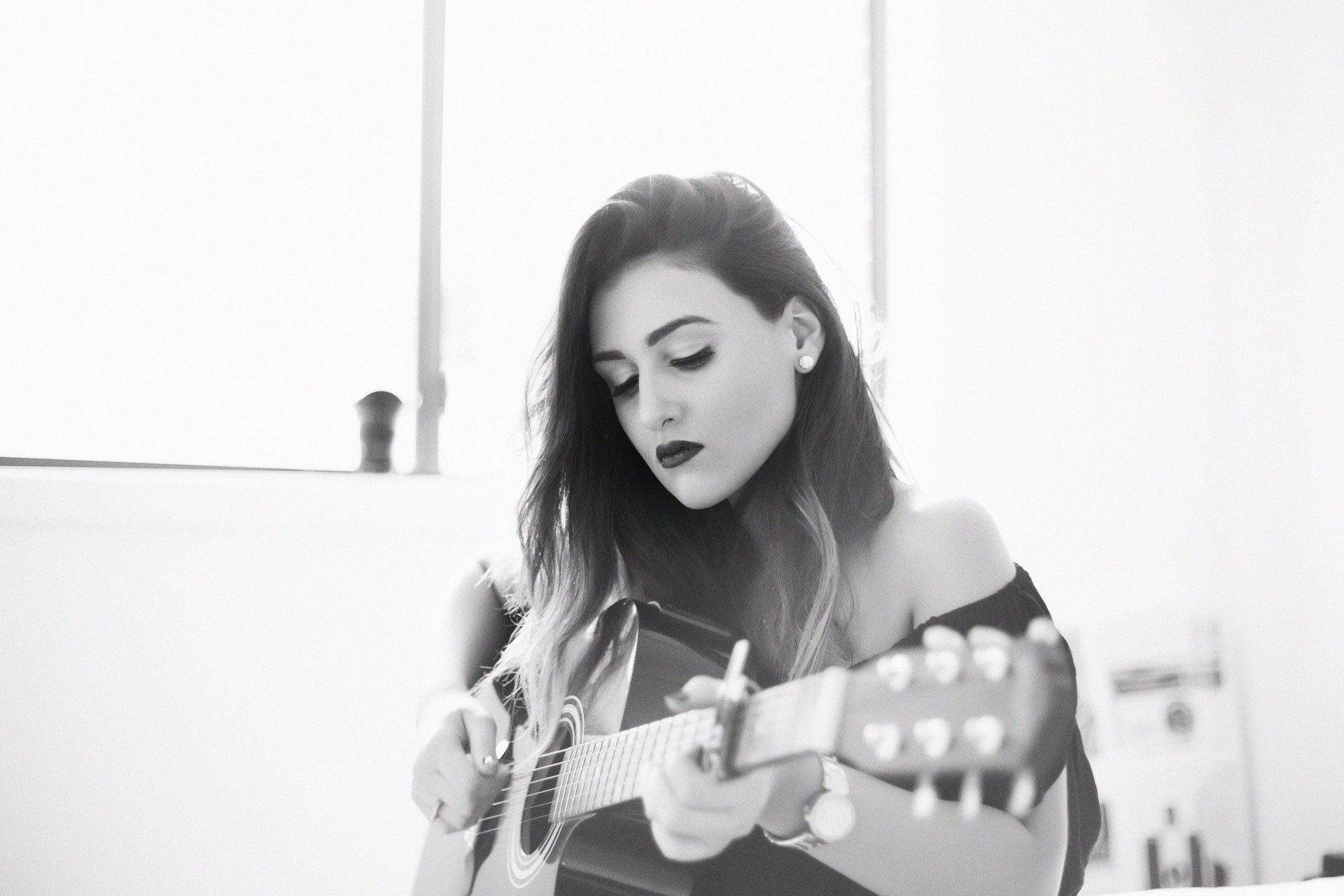 A black-and-white close-up of a person with long hair playing an acoustic guitar while looking down at the instrument.