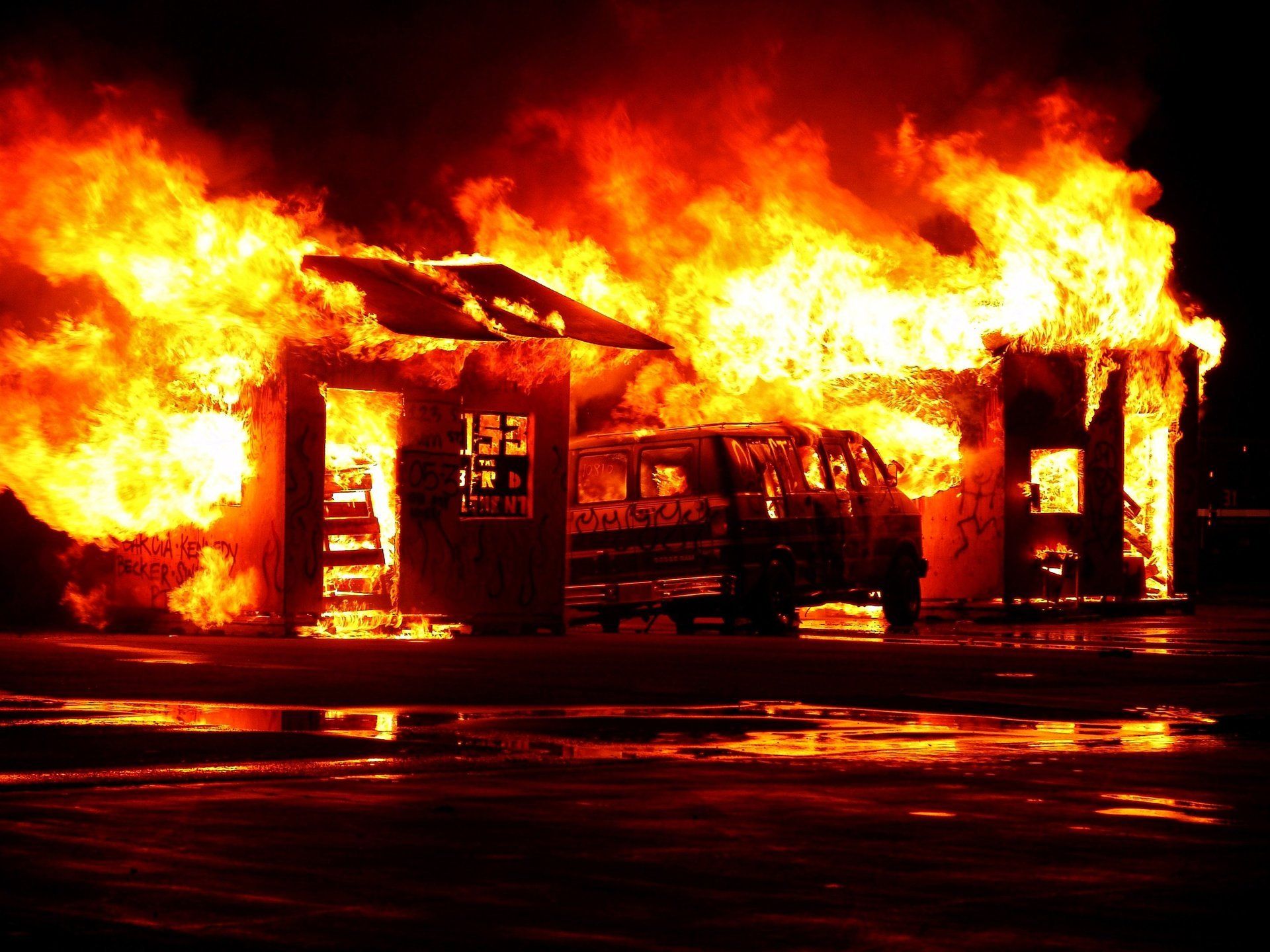 Brennendes Gebäude und Lieferwagen in Flammen, dunkle Nacht, intensives orange-rotes Feuer.