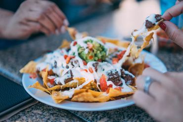 Nachos on a plate with hands reaching for them, cheese, guacamole, sour cream, and tomatoes.