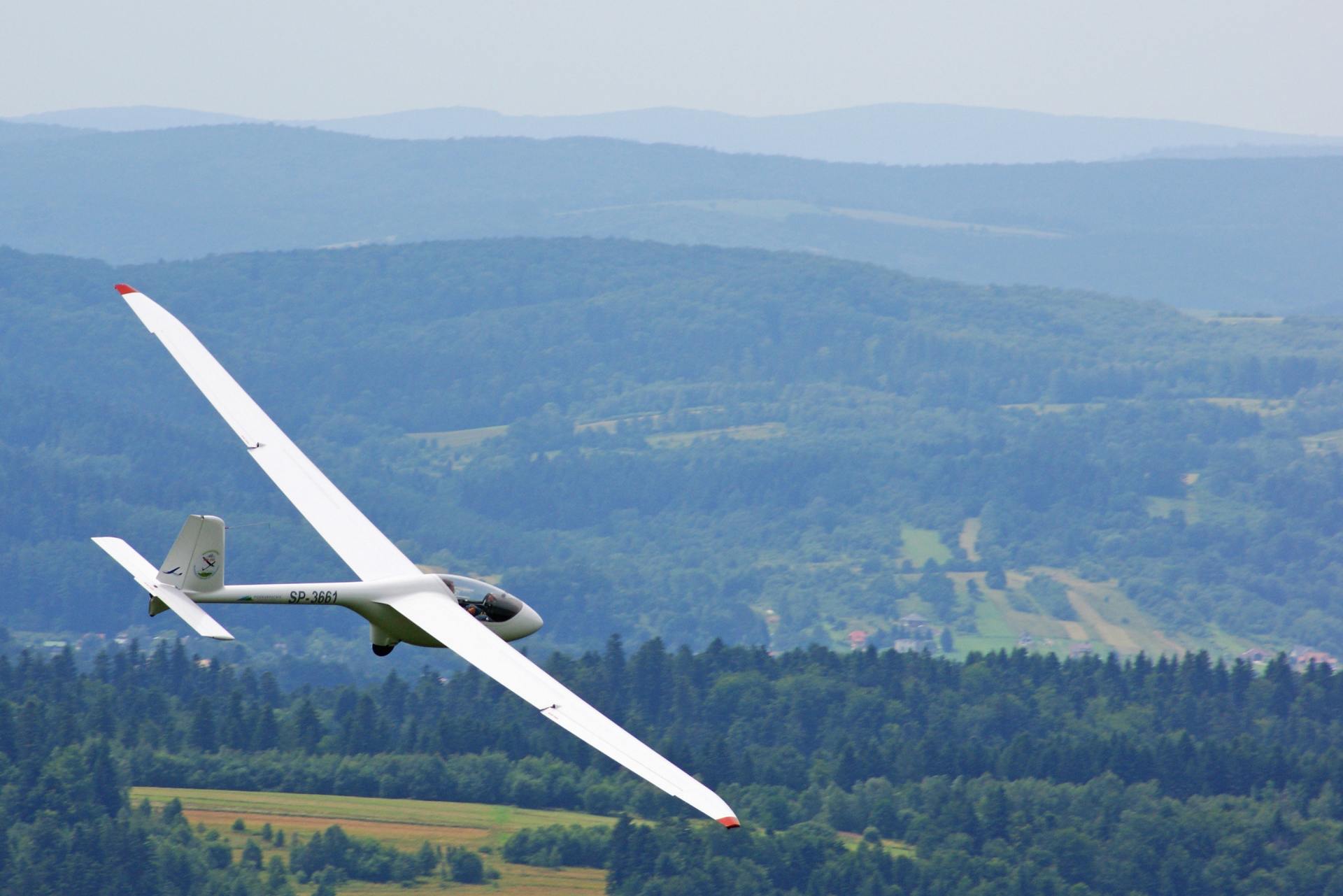 Glider soaring over a forested mountain range on a clear day.