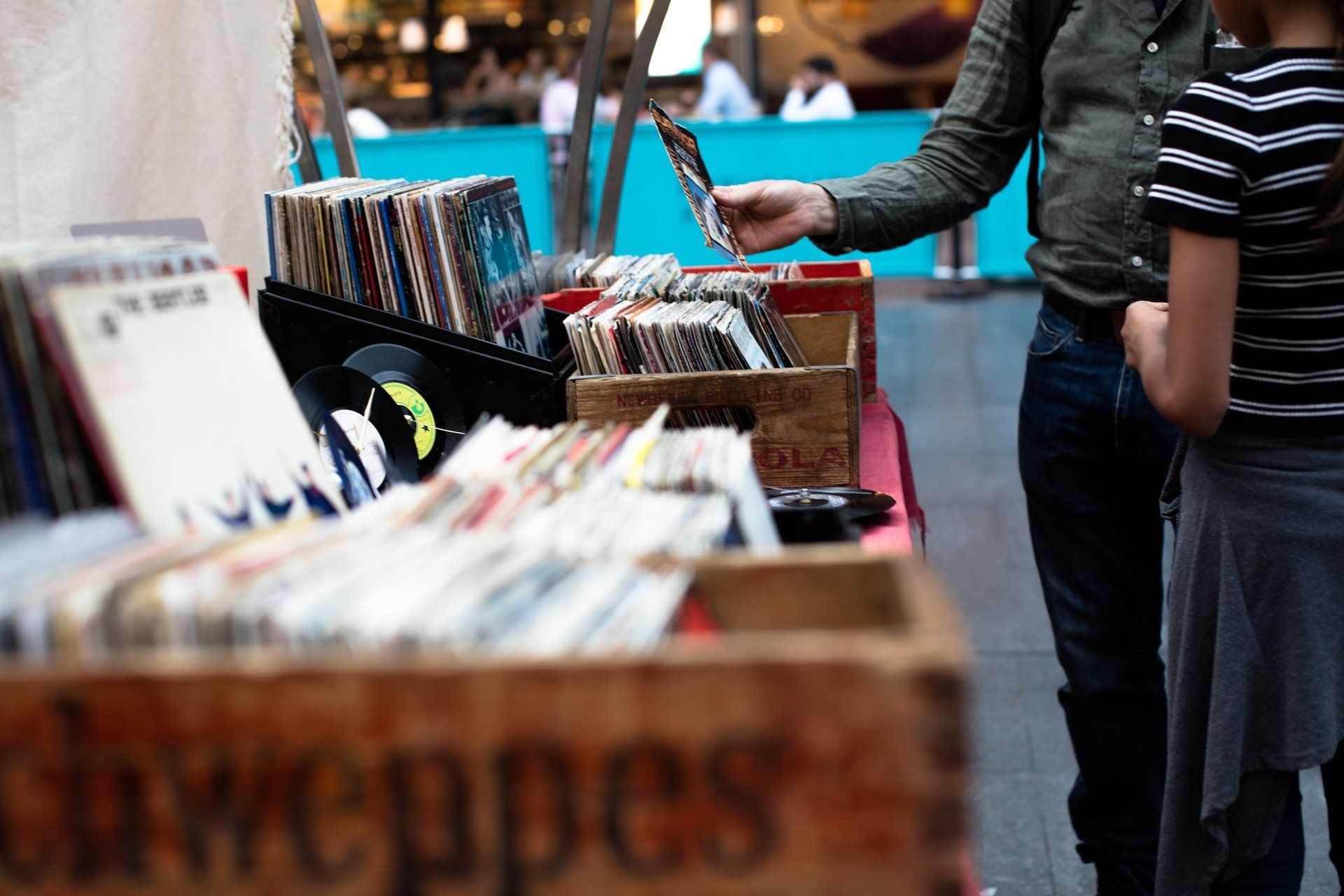 Person browsing records at a vintage shop, with another person observing. Outdoor setting with wooden crates.