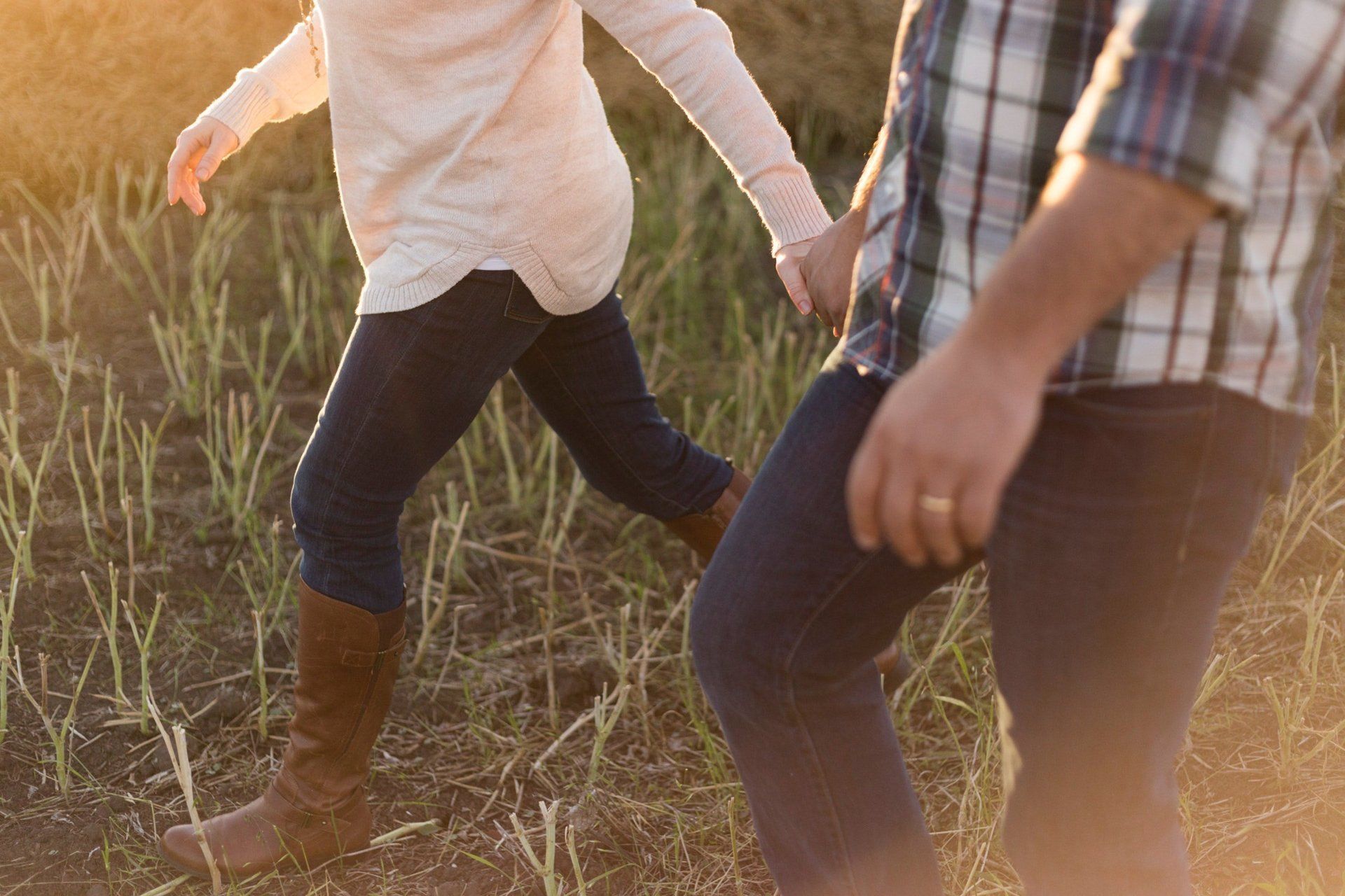 Couple holding hands, walking through tall grass in a field; sunlight casts a warm glow.